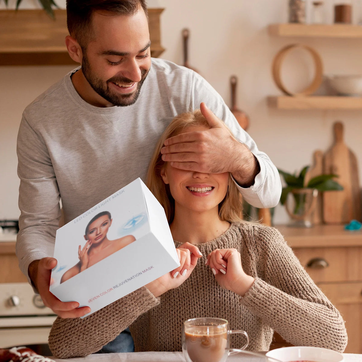 A man surprises a woman with a light therapy face mask gift while she smiles, enhancing her skincare routine.